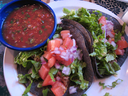 To blue corn tacos stuffed with shredded beef topped with cheese, lettuce, tomatoes and red onions with a bowl of salsa